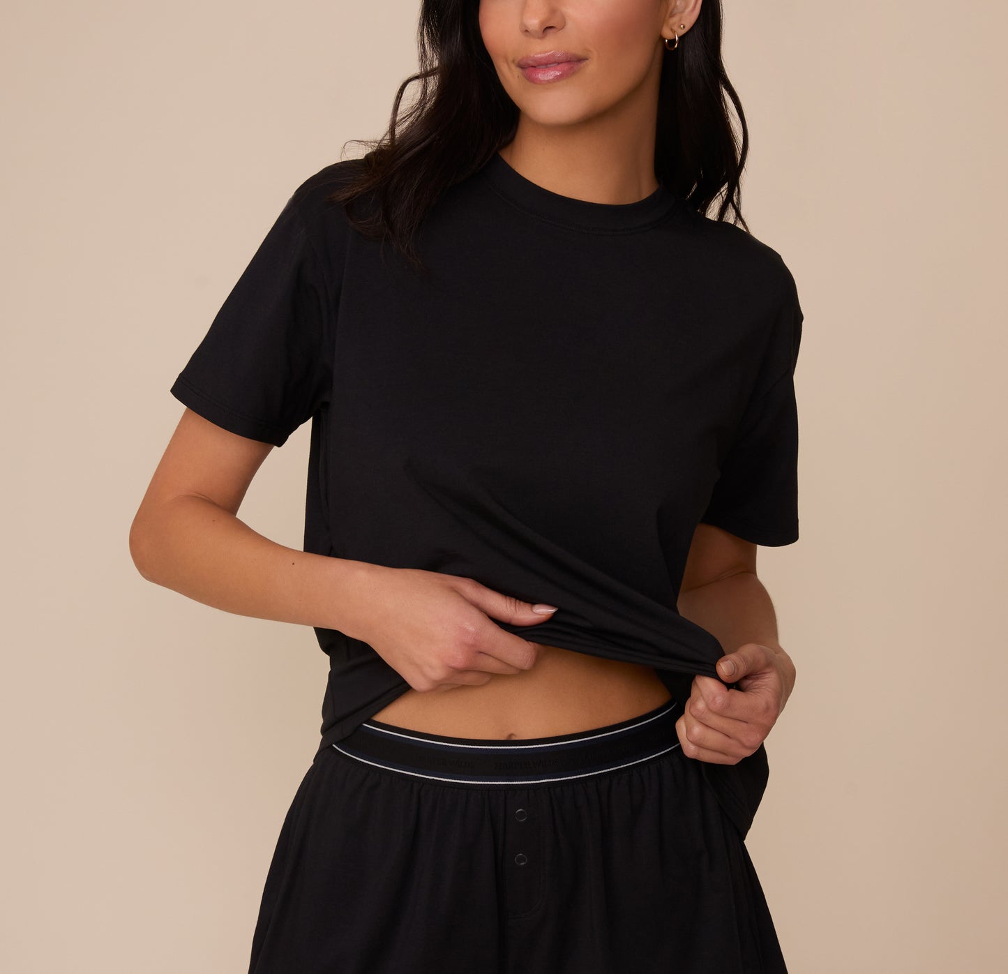 Close-up of Marcela lifting the hem of the Cloud Cotton T-Shirt in Black to reveal the waistband of the matching Cloud Cotton Short. The crewneck tee has a relaxed fit and soft drape, made from breathable cotton-modal fabric. sizing: Marcela wears size XS