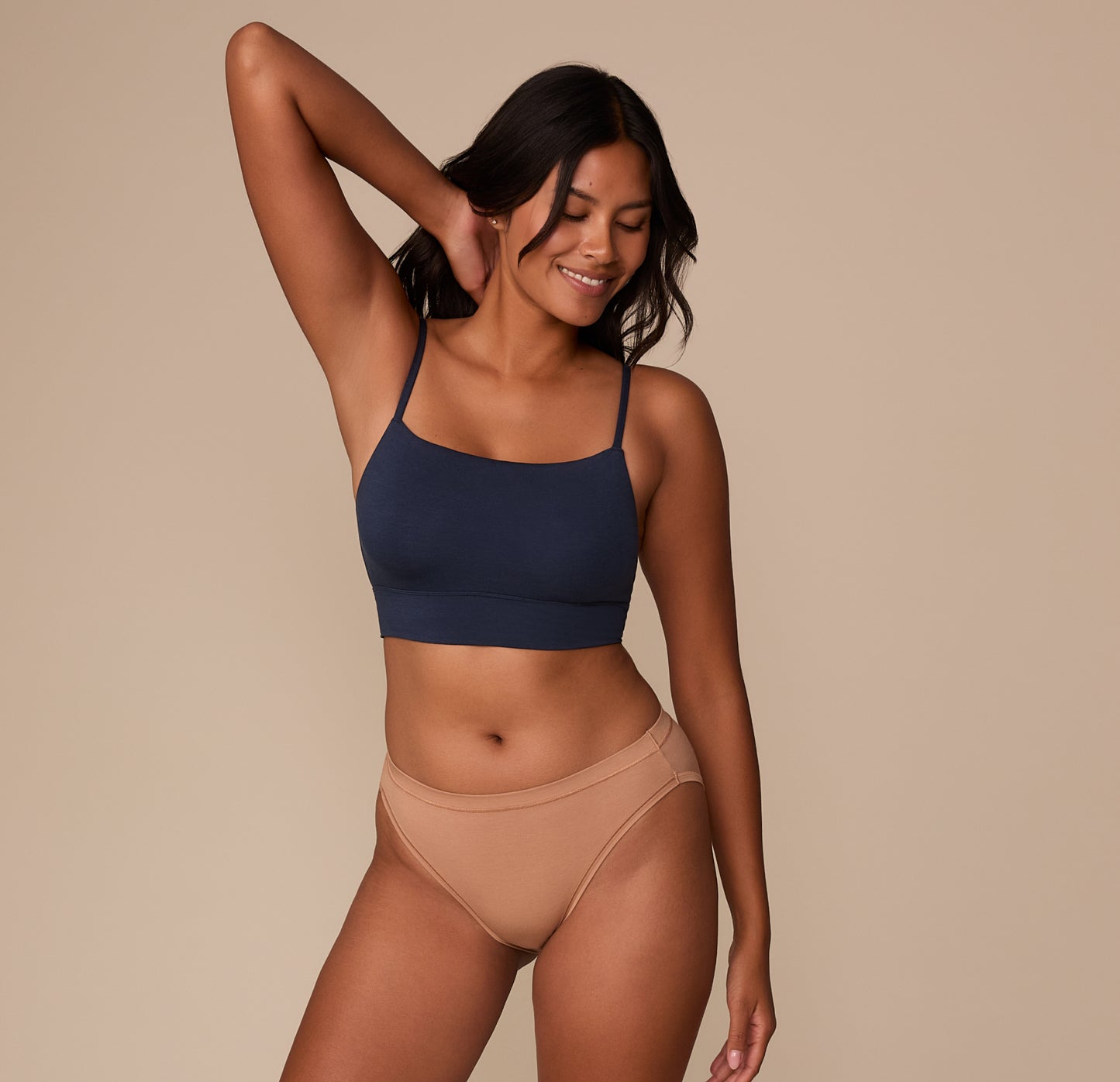 Victoria smiling with one arm raised, wearing the Cloud Cotton Thin Strap Bralette in Midnight and Cloud Cotton French Cut Brief in Dune. The image emphasizes the soft, stretchy fit and everyday comfort.