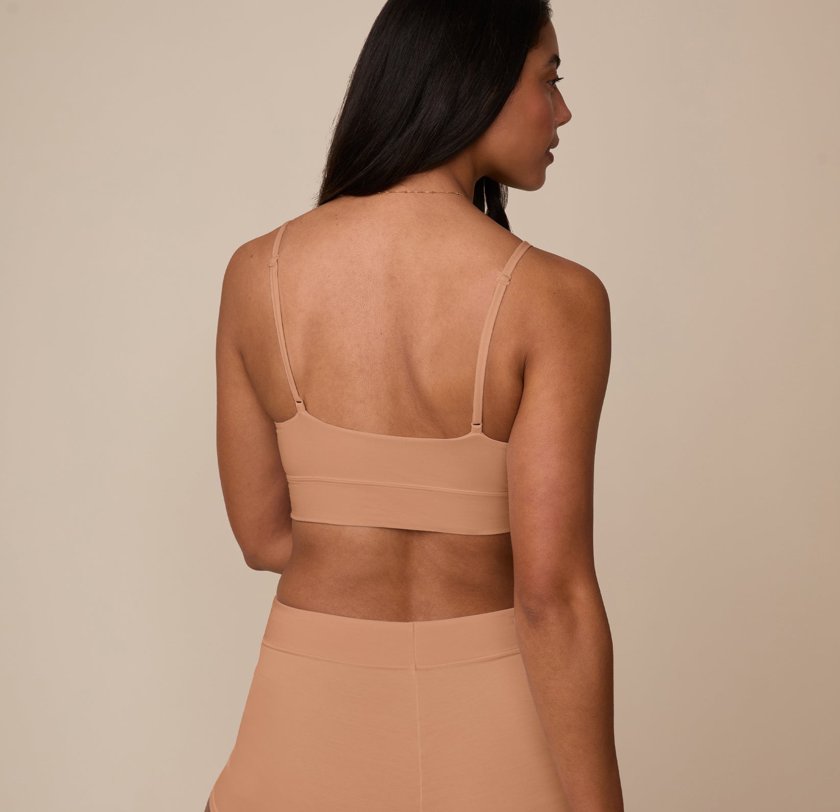Nemesis turned away from the camera wearing the Cloud Cotton Triangle Bralette in Dune. The scoop-back style and ultra-smooth stitching offer a sleek, invisible finish under clothing.