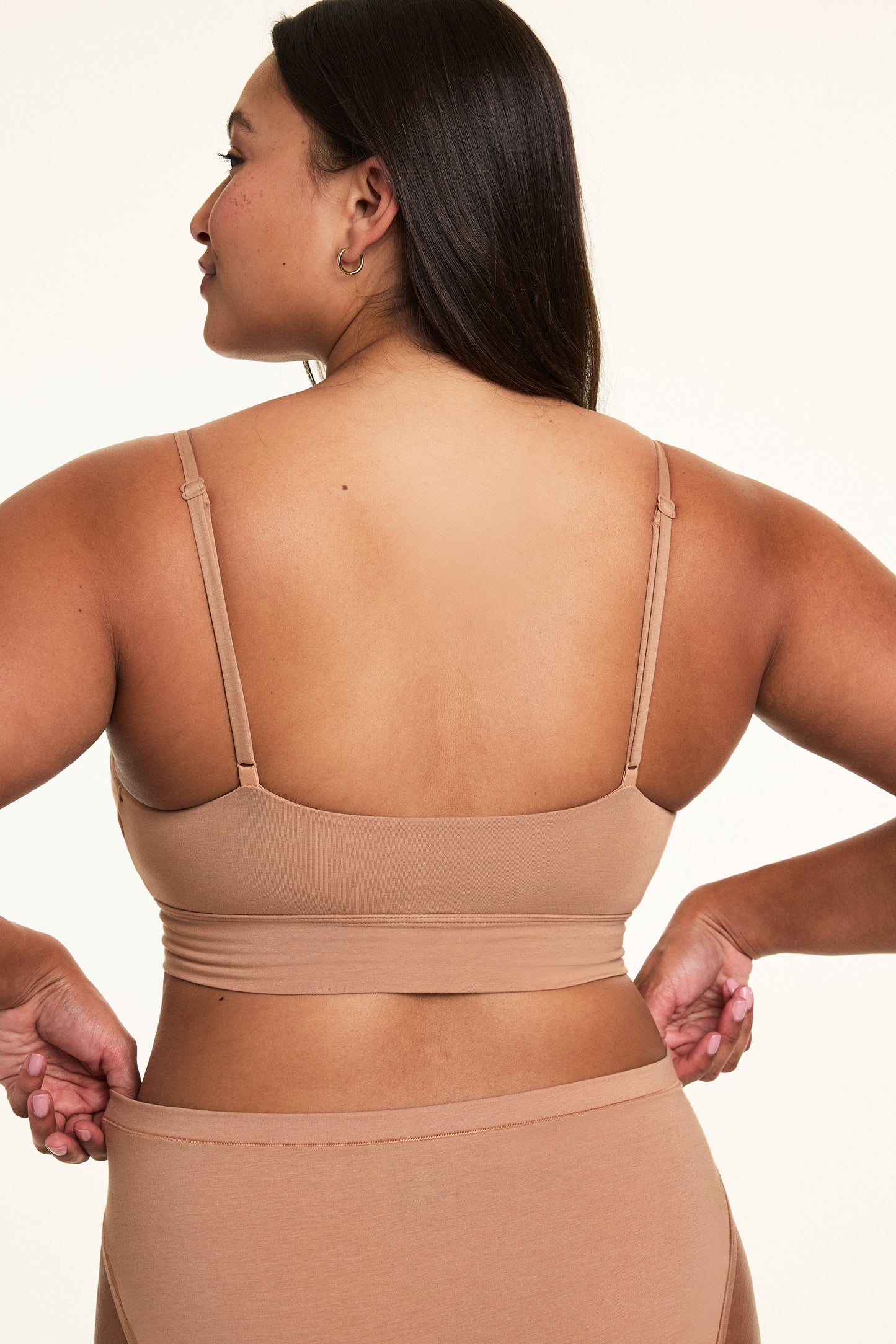 In a softly lit studio, Alana wears the Cloud Cotton Triangle Bralette in Dune with matching Cloud Cotton bottoms, shown from the back. The bralette features thin adjustable straps, a longline band, and soft cotton modal fabric designed for lightweight everyday comfort.