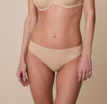 Gracie wearing the No-Show Thong in Beige. The smooth, seamless waistband sits flat against her skin, providing a comfortable and invisible look under clothing. sizing: Gracie wears size XS