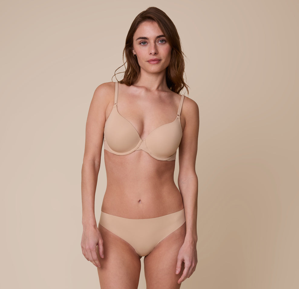 Gracie wearing the No-Show Thong in Beige, paired with a matching Base T-Shirt Bra. She poses with a soft smile, showcasing the thong’s flattering, seamless design.