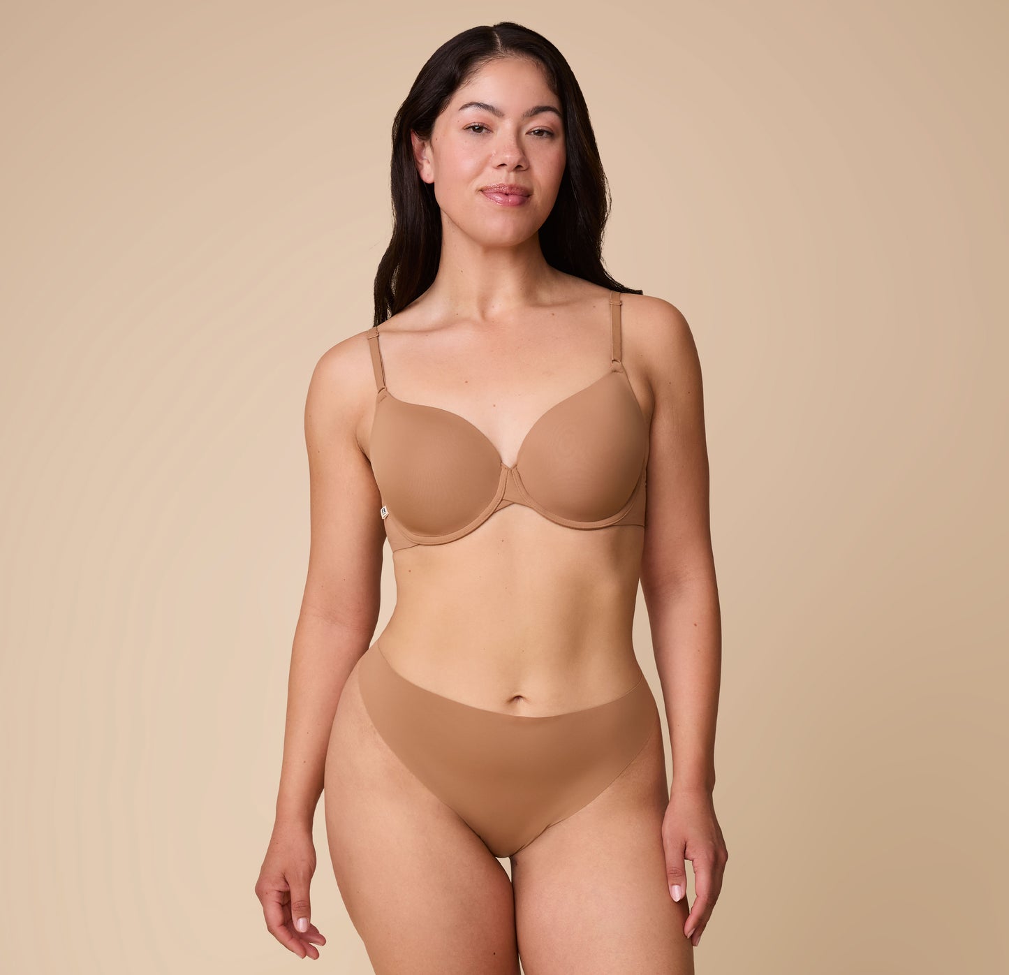 Emersyn standing confidently in the No-Show Thong in Tan, paired with a matching Base T-Shirt Bra, against a neutral backdrop.