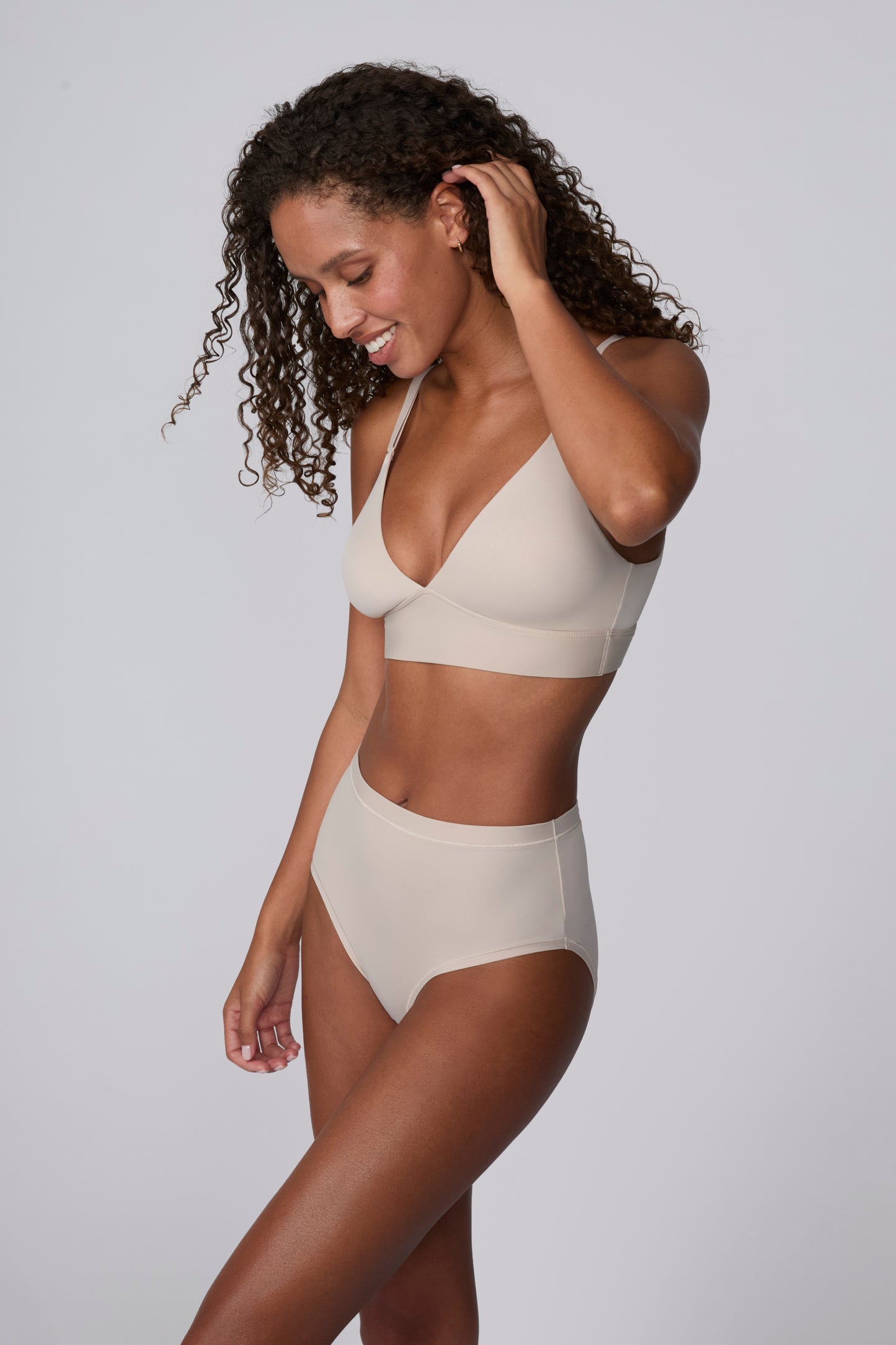 Against a light grey studio backdrop, Courtney wears the Bliss Triangle Bralette in Pebble with matching underwear, looking down with one hand at her hair. The bralette features a deep V neckline, front-adjusting straps, and a wide wireless band in soft microfiber fabric.