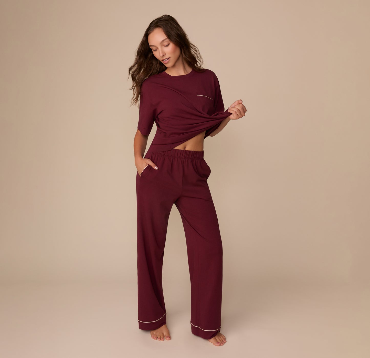 Model Alexis wearing the Perfect Pima Pajama Tee and Pants in Bordeaux, lifting the hem of the tee to show the waistband. The full pant length and pocket design are visible.