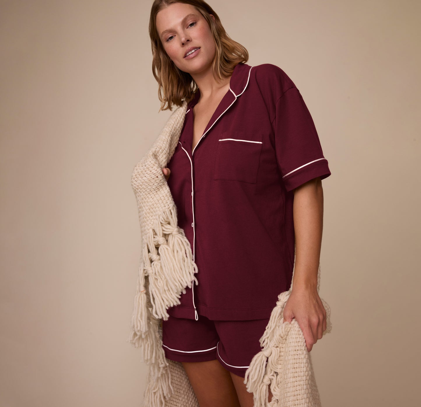 Model Sonnie wearing the Perfect Pima Pajama Shirt and Shorts in Bordeaux, holding a cream knit blanket. The image captures a cozy, relaxed lifestyle moment.
