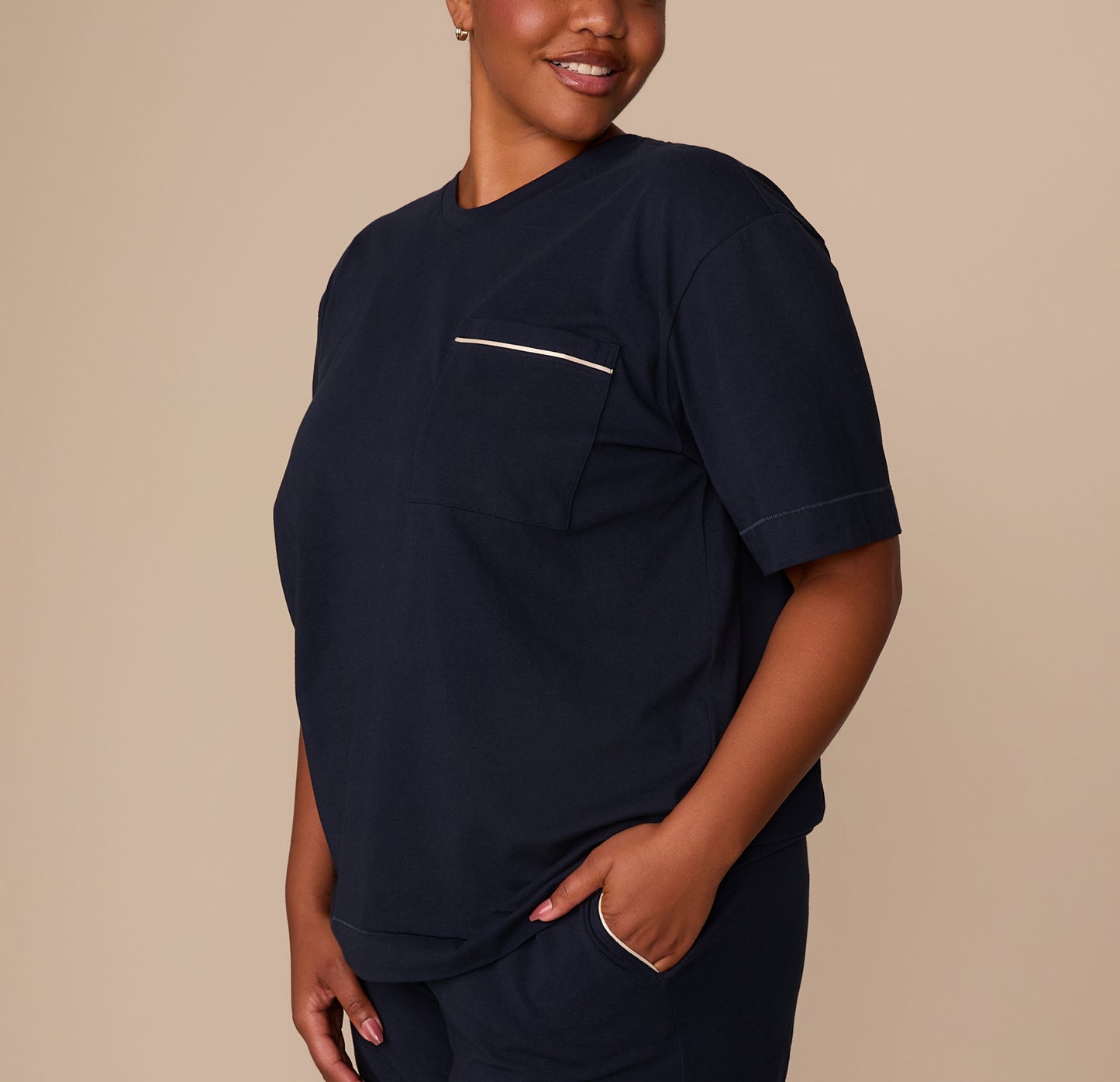 Zarria smiles in a three-quarter profile wearing the navy tee. The contrast piping on the chest pocket is clearly visible, adding subtle definition to the minimalist design.