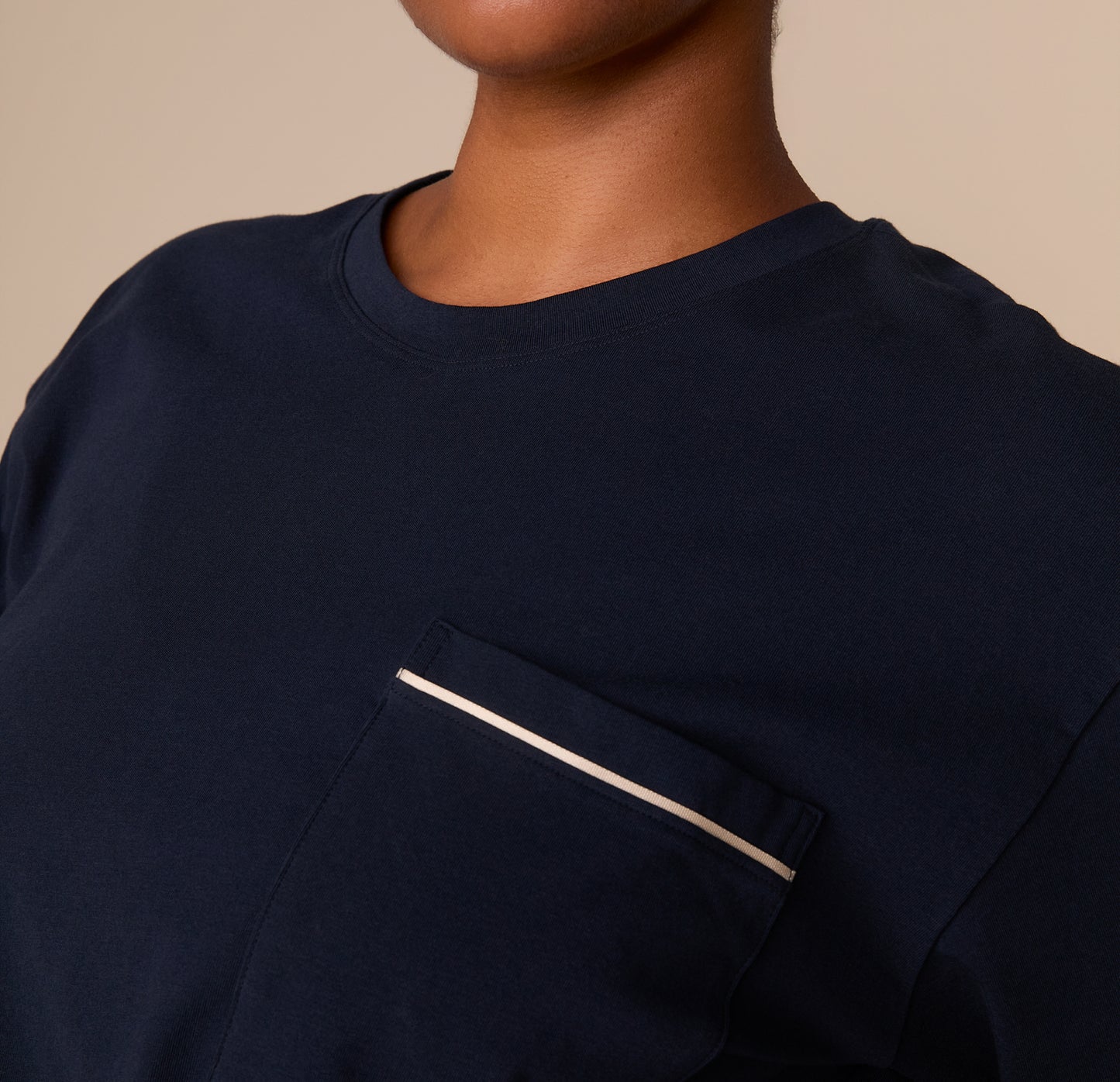 A close-up of the chest pocket reveals fine ivory piping and smooth cotton texture of the navy fabric, showcasing the craftsmanship and elevated finish of the tee.