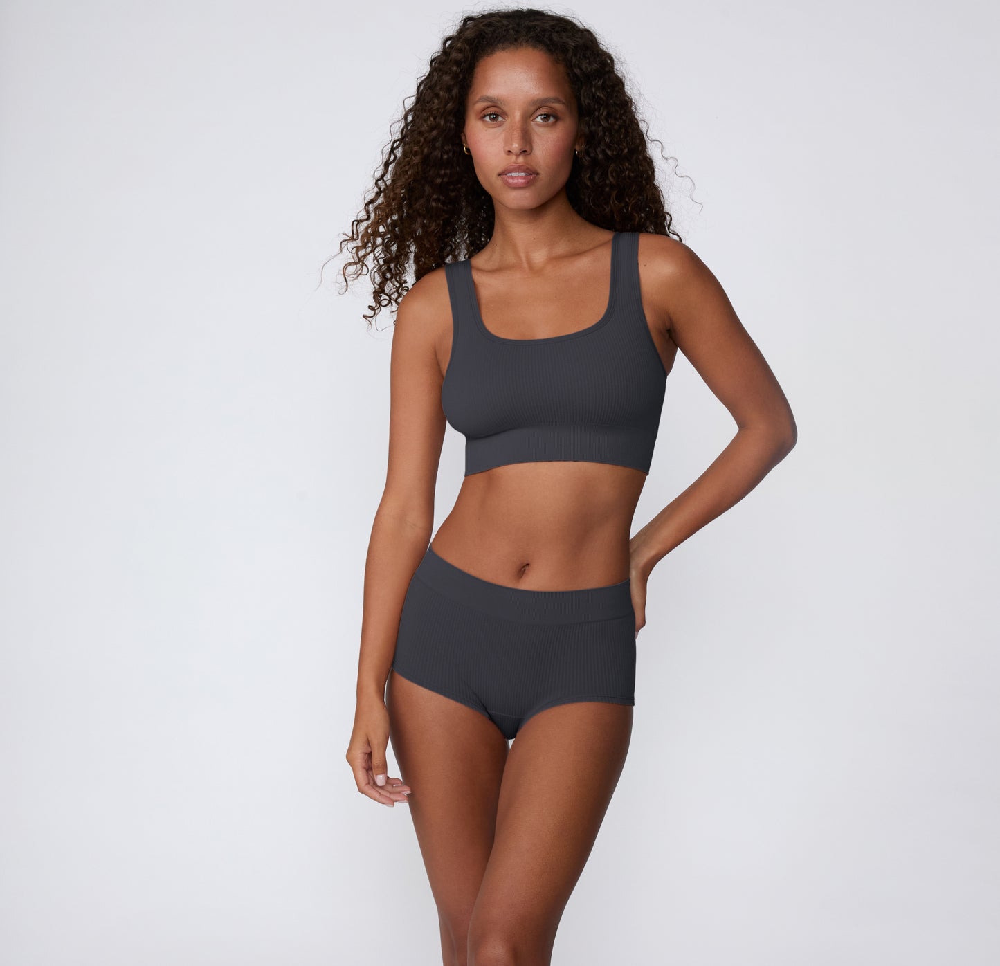 On a light studio background, a model stands wearing the Ribbed Boyshort in Charcoal, paired with a matching ribbed bralette to show the full set.