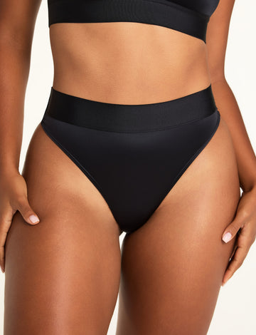 Front view of model wearing the Stretch Satin Thong in Black with a smooth satin front panel and wide satin waistband, paired with the Stretch Satin Square Neck Bralette in Black sizing: Amber wears size Large
