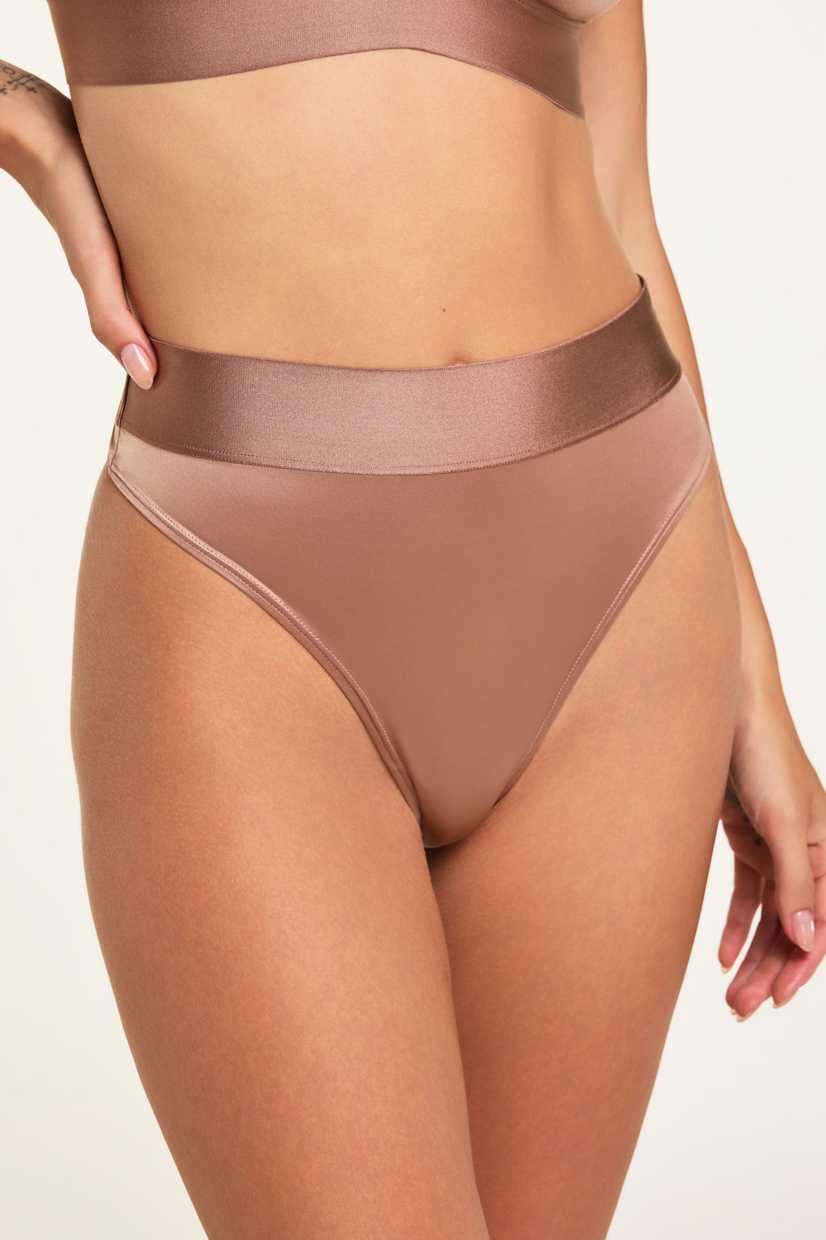 On a light studio backdrop, Holly wears the Stretch Satin Highwaist Thong in Champagne, showing the silky satin fabric, high waist, and wide waistband. sizing: Holly wears size Small