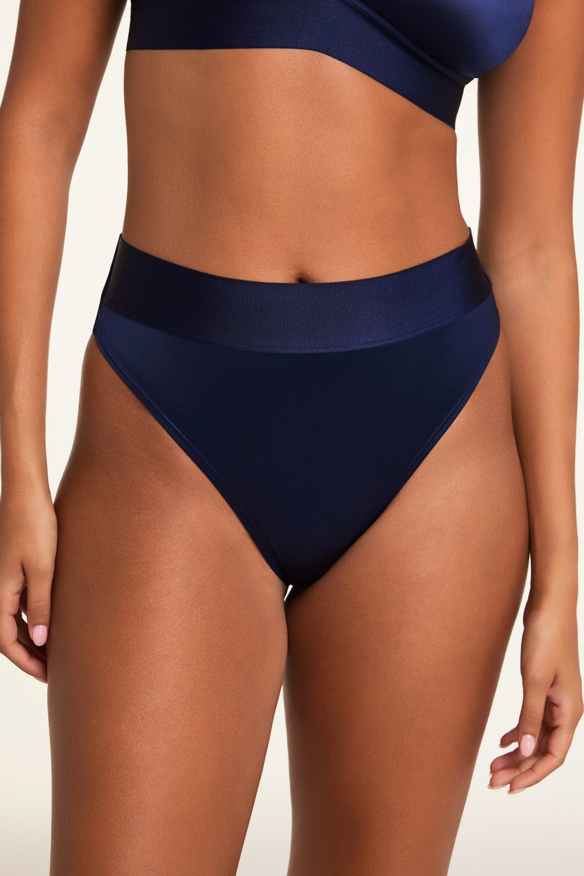 On a light studio backdrop, Kushi wears the Stretch Satin Triangle Bralette and Stretch Satin Highwaist Thong in Navy, shown in a close-up of the high-waisted thong and wide top band. sizing: Kuhsi wears size Small