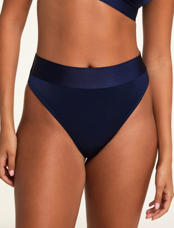 On a light studio backdrop, Kushi wears the Stretch Satin Triangle Bralette and Stretch Satin Highwaist Thong in Navy, shown in a close-up of the high-waisted thong and wide top band. sizing: Kuhsi wears size Small