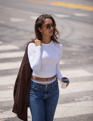 On a city street, Laura wears the Bliss Long-Sleeve Tee in White with the Bliss French-Cut Brief in Beige layered under blue jeans, carrying a coffee cup and brown coat. sizing: Kushi wears size Small 