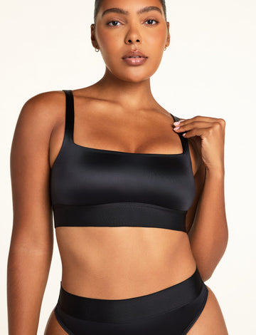 On a warm white studio backdrop, Amber wears the Stretch Satin Square Neck Bralette in Black and Stretch Satin Highwaist Thong in Black, showing the square neckline, thin straps, longline band, and wide thong waistband. sizing: Amber wears size Medium