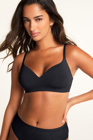 Against a warm white studio backdrop, a model looks toward the camera wearing a black V-neck bra with adjustable straps and black underwear with lace detailing at the hips. sizing: Kushi wears size Small