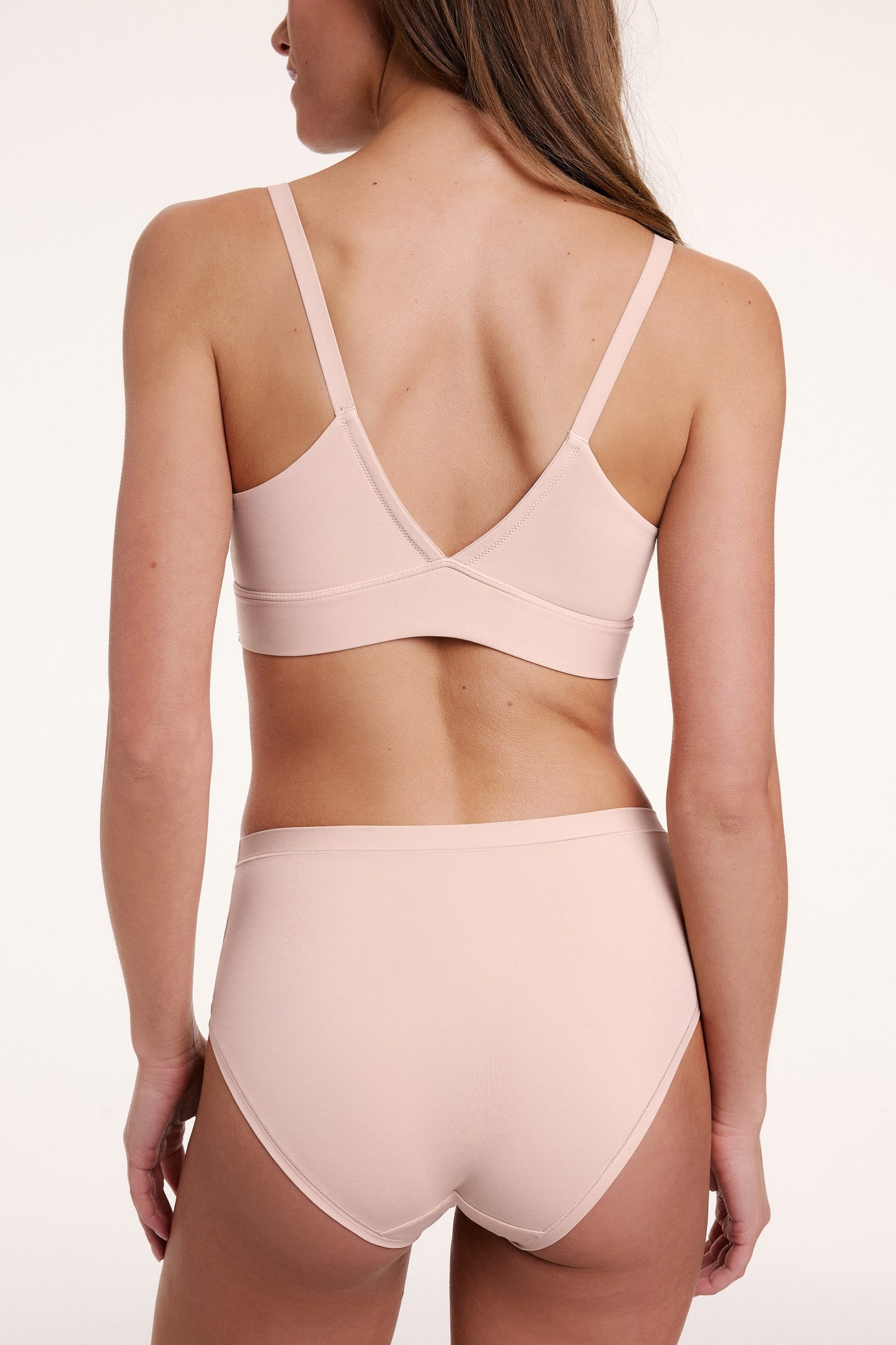 On a light studio backdrop, Gianna wears the Bliss Triangle Bralette in Rose, shown from the back. The bralette features a V-shaped back, adjustable straps, and a smooth underband.