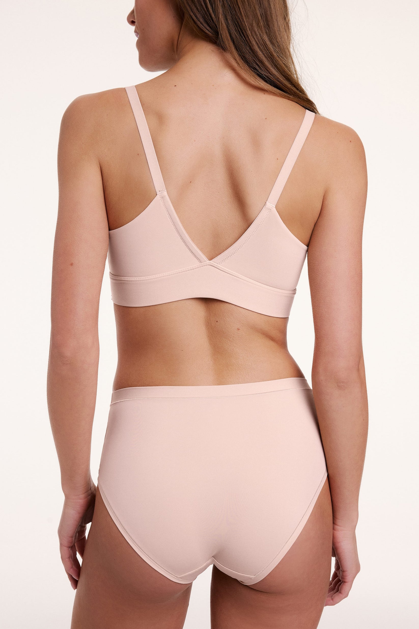 On a light studio backdrop, Gianna wears the Bliss Triangle Bralette in Rose, shown from the back. The bralette features a V-shaped back, adjustable straps, and a smooth underband.