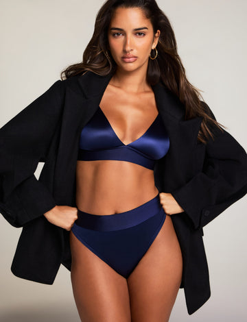 In a studio setting, Kushi wears the Stretch Satin Triangle Bralette and Stretch Satin Highwaist Thong in Navy, layered under a black blazer. The bralette features a deep V-neck and adjustable strap