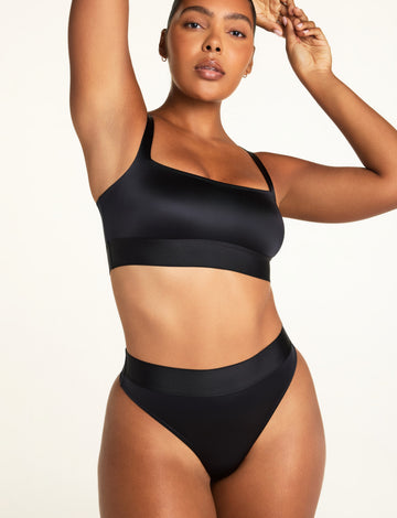 Front angle view of the Stretch Satin Thong in Black on model, highlighting the smooth satin front panel, high-cut leg opening, and wide waistband, paired with the Stretch Satin Square Neck Bralette in Black
