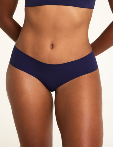 In a light studio backdrop, Amber wears the No-Show Hiphugger in Noir, shown in close-up with its smooth, raw-edge finish; the bottom of the WhippedKnit V-Neck in Noir is visible at the top of the frame. sizing: Amber wears size Large 