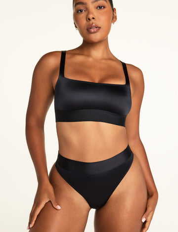 On a warm white studio backdrop, Amber wears the Stretch Satin Square Neck Bralette in Black and Stretch Satin Highwaist Thong in Black, facing the camera to show the square neckline, smooth satin fabric, and longline silhouette.