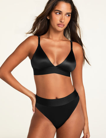 On a light studio backdrop, Kushi wears the Stretch Satin Triangle Bralette and Stretch Satin Highwaist Thong in Black, showing the deep V-neck bralette and high-waisted thong with a wide top band.