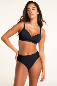 A model poses against a warm white studio backdrop wearing a black bra with a V-neckline and adjustable straps, paired with matching black underwear featuring lace panels on both sides.