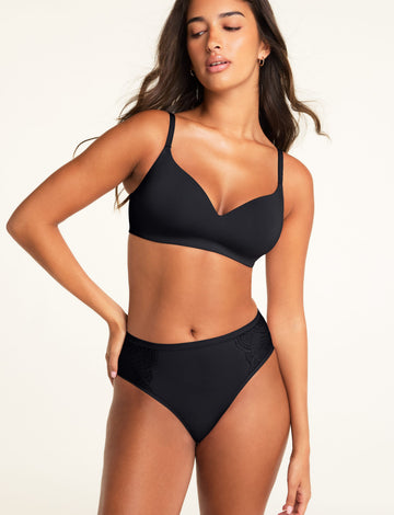 A model poses against a warm white studio backdrop wearing a black bra with a V-neckline and adjustable straps, paired with matching black underwear featuring lace panels on both sides.