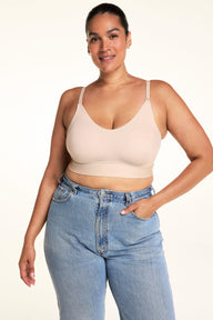 Jennifer smiles while looking down in a three-quarter view, wearing the Bliss Nursing Bralette in Beige with light-wash jeans. The angle shows the scoop neckline, smooth cups, adjustable straps, and wide underband.