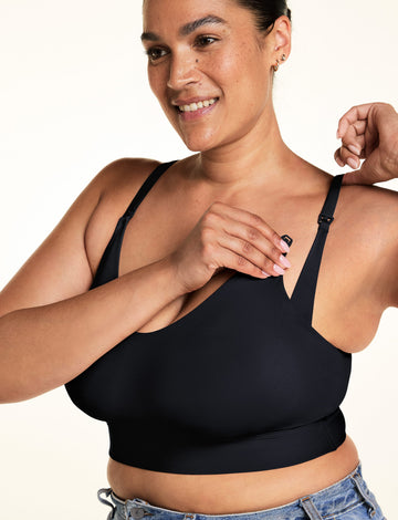 Jennifer demonstrates the nursing clip on the Black Bliss Nursing Bralette, holding the front strap clasp while lifting the shoulder strap. The close-up highlights the adjustable straps and nursing-access detail against a warm white studio backdrop.