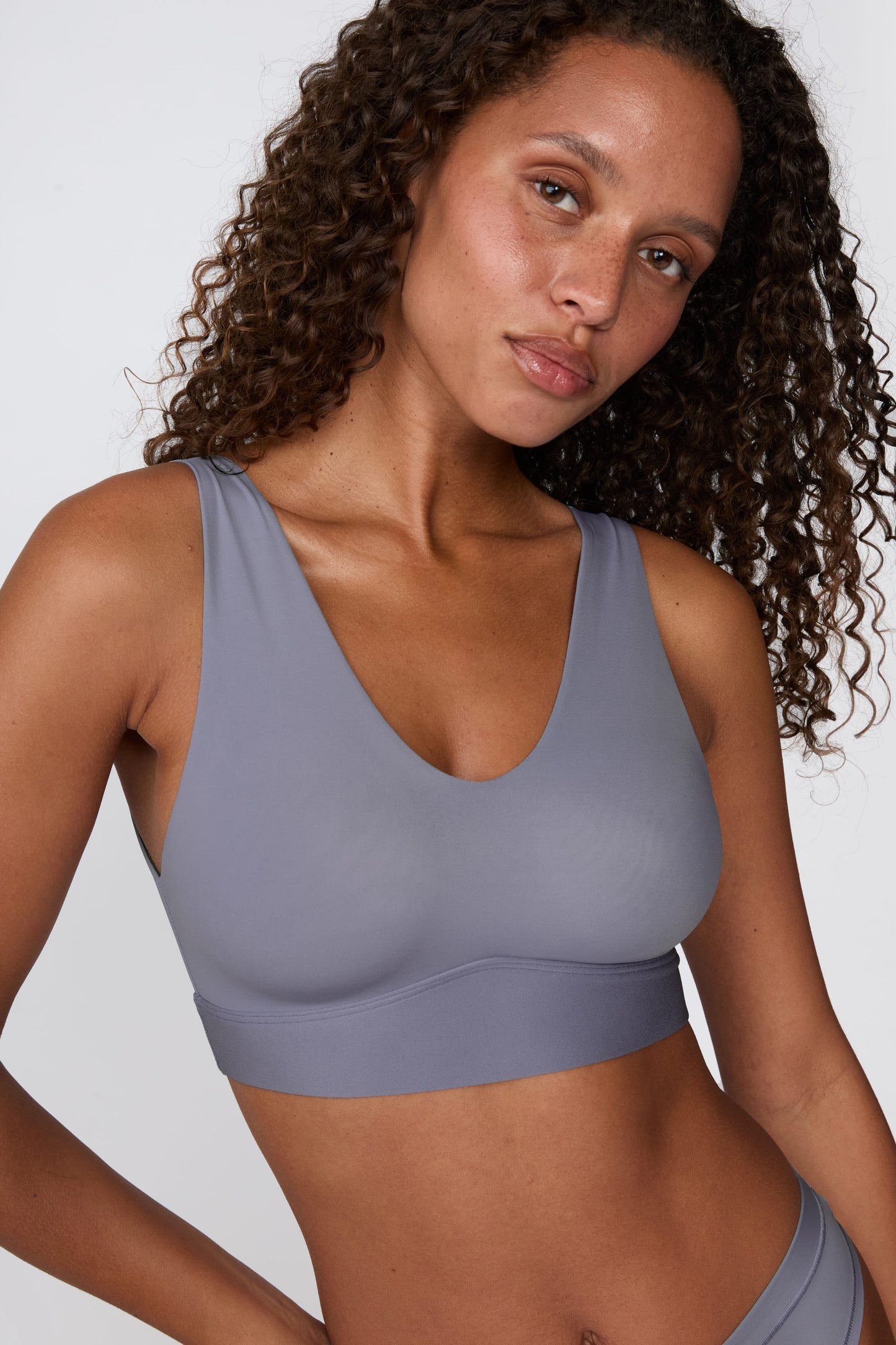 On a white studio backdrop, Courtney wears the Bliss Bralette in Steel, shown in a close-up. The bralette features a deep V-neckline, wide straps, and smooth, lightweight fabric.