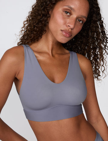 On a white studio backdrop, Courtney wears the Bliss Bralette in Steel, shown in a close-up. The bralette features a deep V-neckline, wide straps, and smooth, lightweight fabric.
