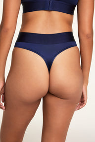 On a light studio backdrop, Kushi wears the Stretch Satin Highwaist Thong in Navy, shown from the back with its wide waistband and satin finish.