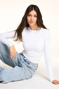 On a light studio backdrop, Laura wears the Bliss Long-Sleeve Tee in White with the Bliss French-Cut Brief in Beige, seated in light-wash jeans. sizing: Laura wears size Small 
