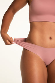 On a white studio backdrop, Amber wears the Whipped Knit Scoop and No-Show Low-Rise Thong in Plum, gently stretching the thong waistband to show its flexibility.