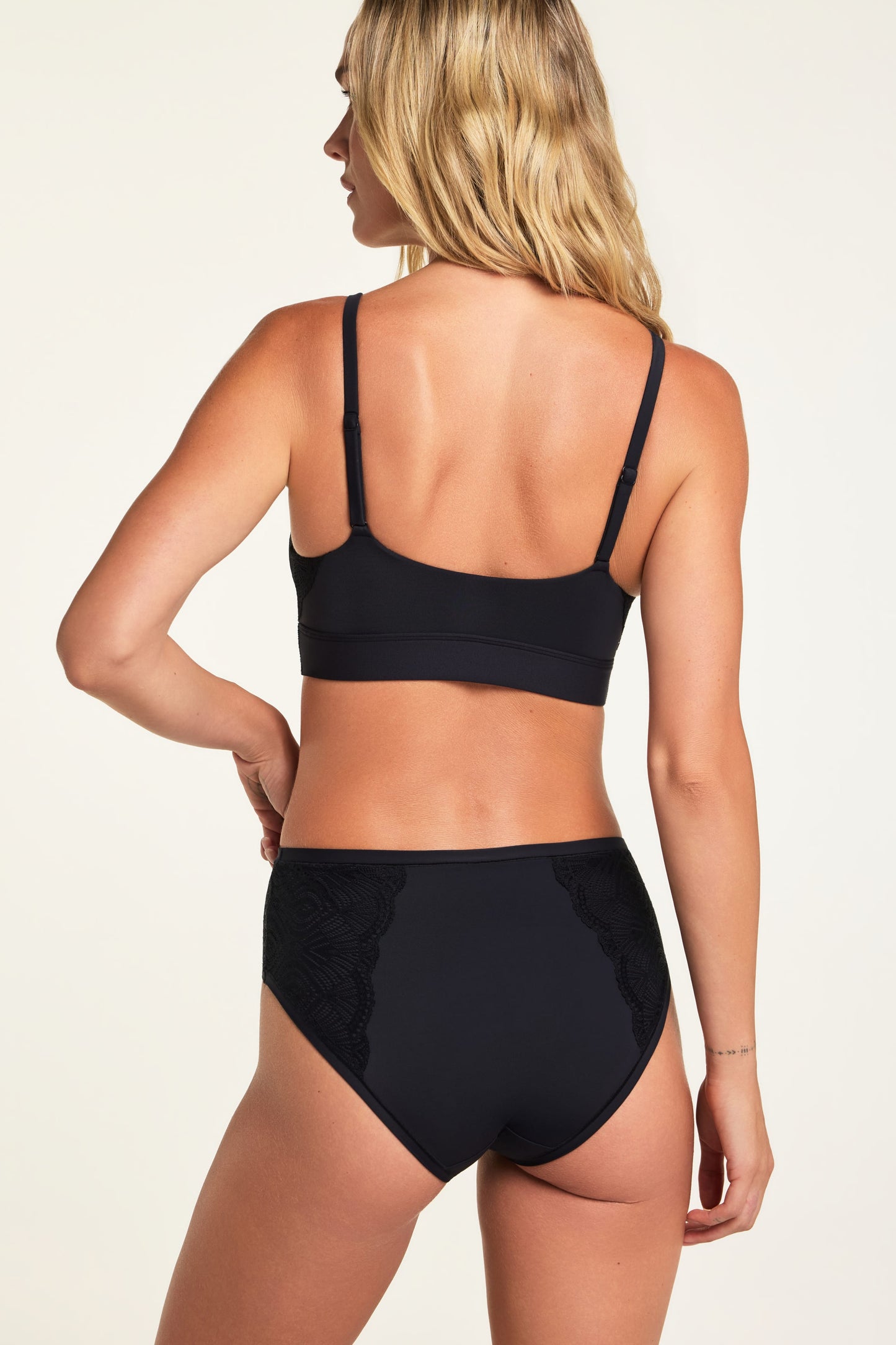 On a warm white studio backdrop, Holly wears the Bliss Lace Bralette in Black and Bliss Lace Highwaist Brief in Black, shown from the back to highlight the smooth bralette back, adjustable straps, and lace panels on the brief.