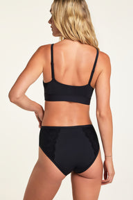 On a warm white studio backdrop, Holly wears the Bliss Lace Bralette in Black and Bliss Lace Highwaist Brief in Black, shown from the back to highlight the smooth bralette back, adjustable straps, and lace panels on the brief.