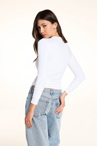On a light studio backdrop, Laura looks over her shoulder wearing the Bliss Long-Sleeve Tee in White with the Bliss French-Cut Brief in Beige and light-wash jeans.
