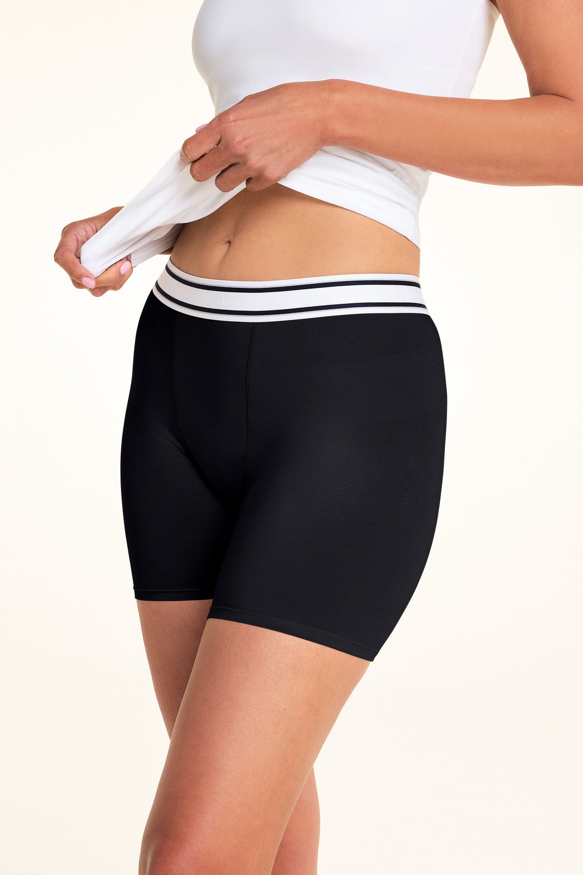 A side-angle close-up of Meagan wearing the Cloud Cotton Boxer Brief in Black with a white camisole. She lifts the camisole to reveal the striped waistband and the smooth, fitted design of the boxer briefs.