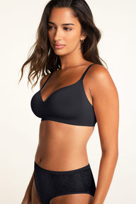 In a studio against a warm white backdrop, Kushi wears the Base Wirefree T-Shirt Bra in Black with the Bliss Lace Highwaist Brief in Black, showing the bra’s V-neckline and smooth cups alongside the brief’s decorative lace side panels.