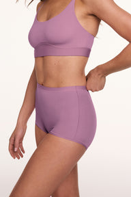 In a studio setting, a model wears the Bliss Boyshort in Mirage with a matching bra, shown in a side view while adjusting the waistband. The boyshort features a mid-rise fit, full coverage, and smooth, seamless fabric. sizing: Gianna wears size Small
