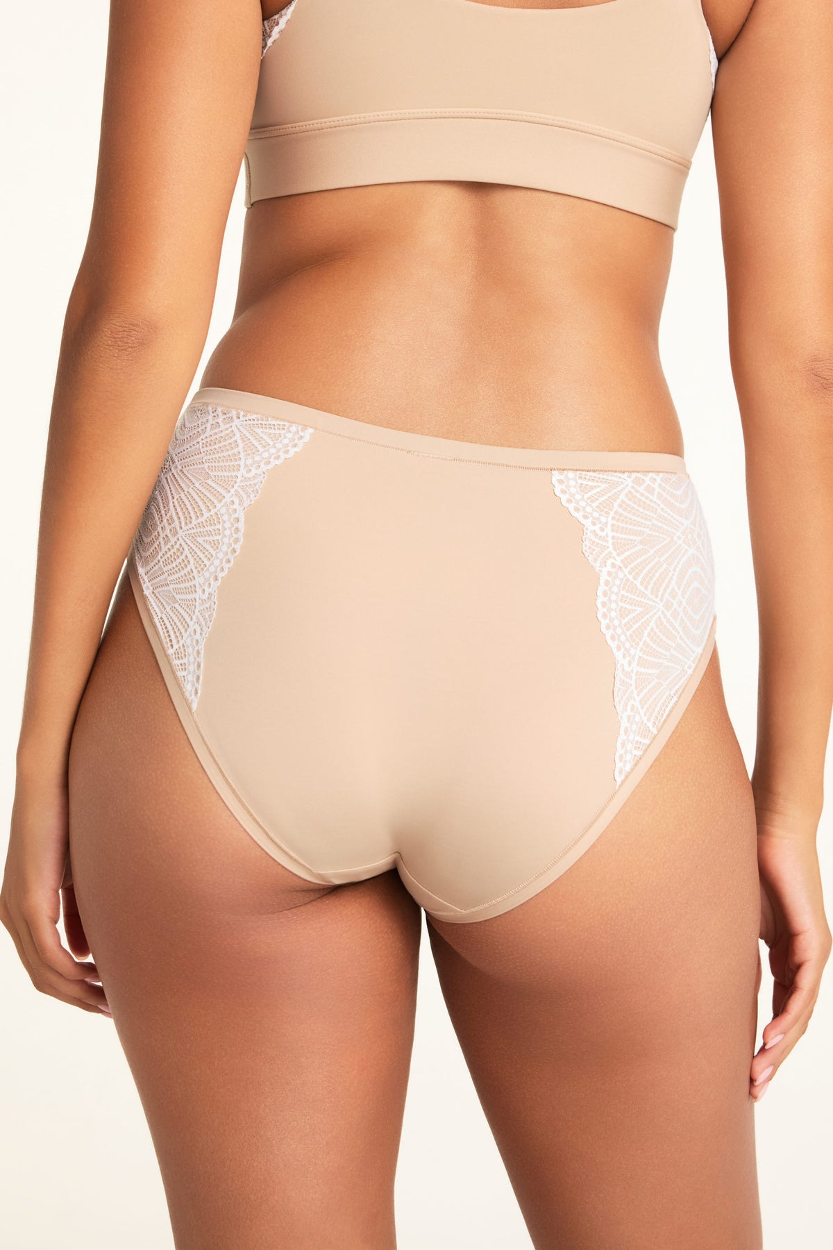 In a bright studio, Kushi wears the Bliss Lace Bralette and Bliss Lace Highwaist Brief in Beige, shown from the back to highlight the lace detailing on the bralette and brief.