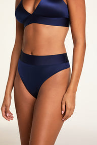 On a light studio backdrop, Kushi wears the Stretch Satin Triangle Bralette and Stretch Satin Highwaist Thong in Navy, shown from a three-quarter angle to highlight the bralette’s deep V-neck and the thong’s high waist and wide top band.