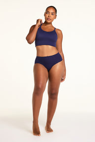 On a warm white studio backdrop, Amber wears the Whipped Knit V-Neck and No-Show Highwaist Brief in Noir in a full-length view, showing the coordinated set.