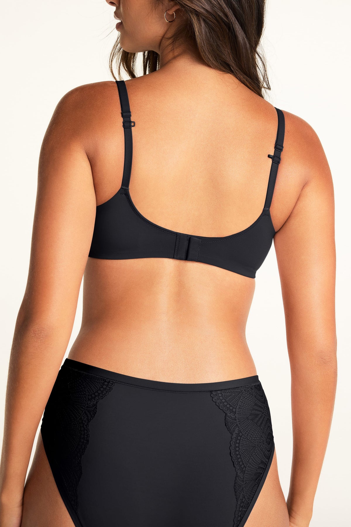 Against a warm white studio backdrop, Kushi shows the back of the Base Wirefree T-Shirt Bra in Black, featuring adjustable shoulder straps and a hook-and-eye closure, paired with the Bliss Lace Highwaist Brief in Black with lace panels at the sides.