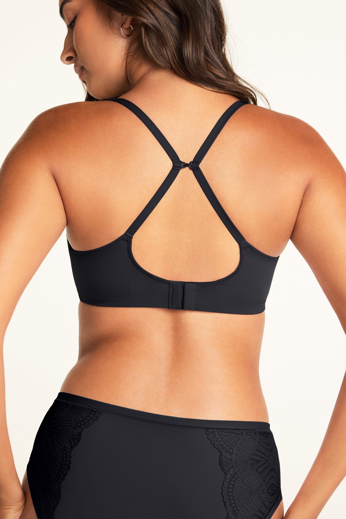 Against a warm white studio backdrop, Kushi models the Base Wirefree T-Shirt Bra in Black with its shoulder straps connected in a cross-back style, paired with the Bliss Lace Highwaist Brief in Black, featuring lace detailing on the si