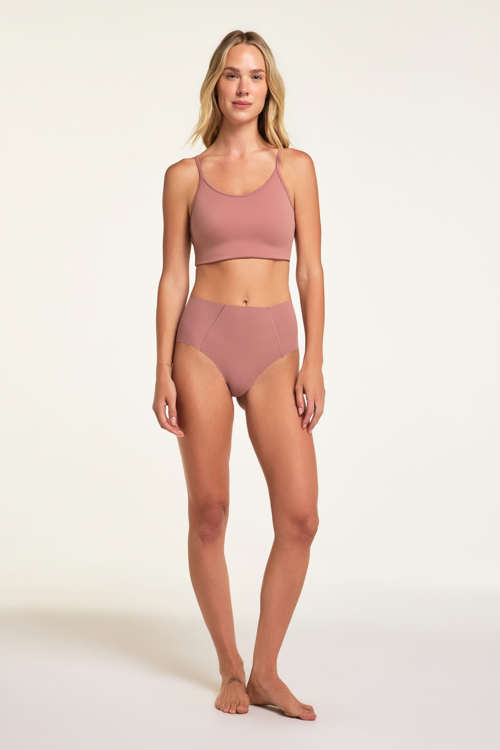 On a white studio backdrop, Holly wears the No-Show Highwaist Brief in Plum with a matching cropped top, showing the full high-waisted brief and angled front seams.