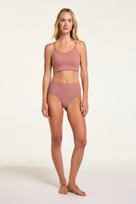 On a white studio backdrop, Holly wears the No-Show Highwaist Brief in Plum with a matching cropped top, showing the full high-waisted brief and angled front seams.