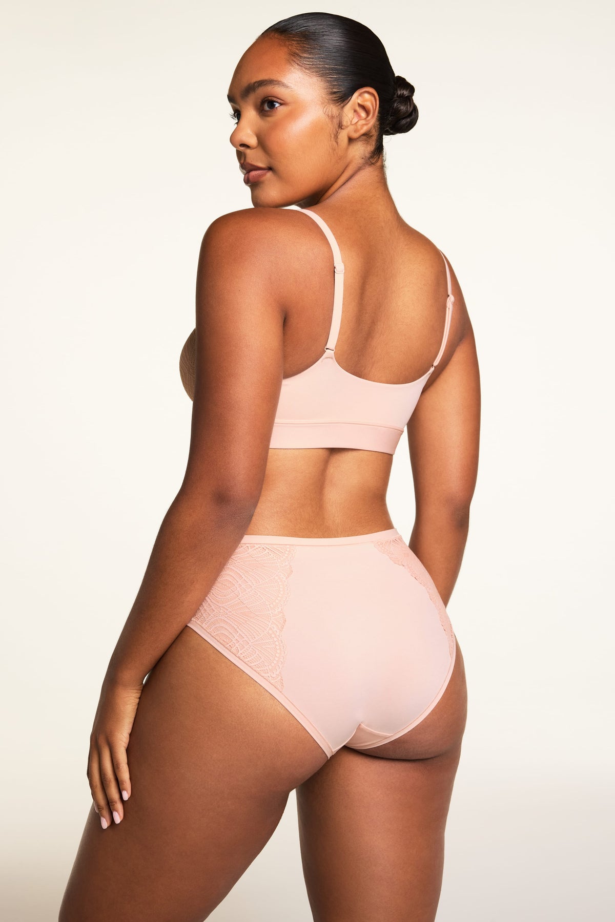 On a warm white studio backdrop, Amber wears the Bliss Lace Bralette in Ember Rose and Bliss Lace Highwaist Brief in Ember Rose, shown from the back to highlight the bralette’s V-back, adjustable straps, smooth back, and lace panels on the brief.